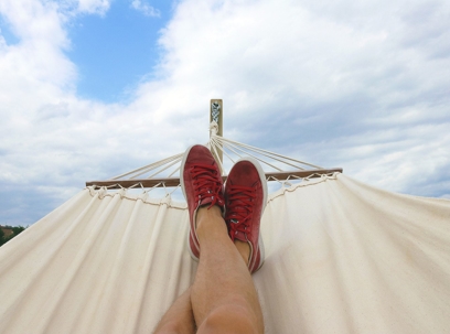 Point of view from a person sitting in a white hammock looking up at their feet. Beyond that is a blue sky with clouds.